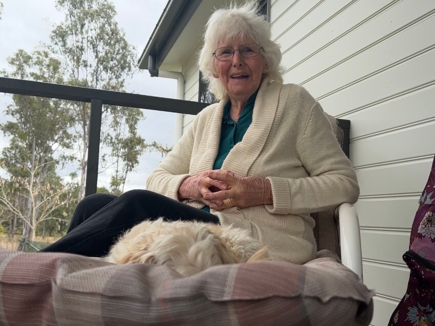 An elderly woman sits on her front porch with her fluffly dog on her chair.