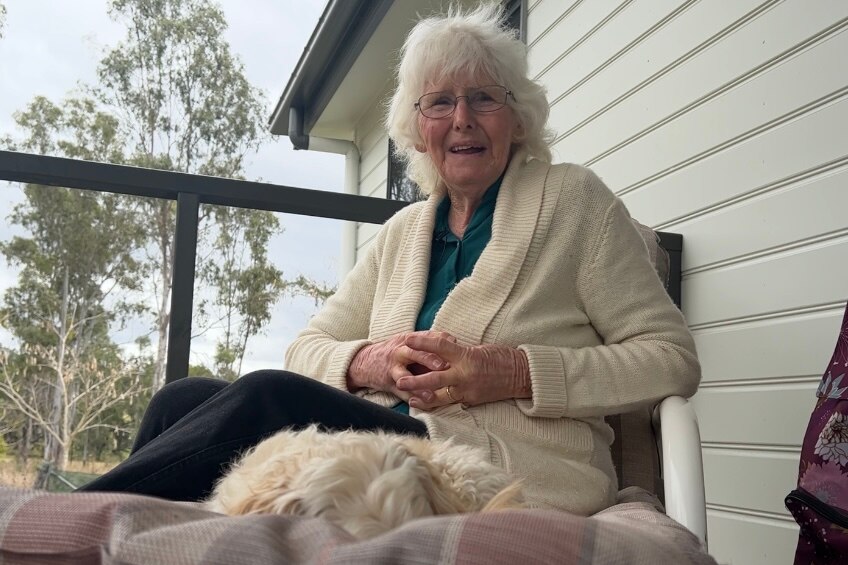 An elderly woman sits on her front porch with her fluffly dog on her chair.