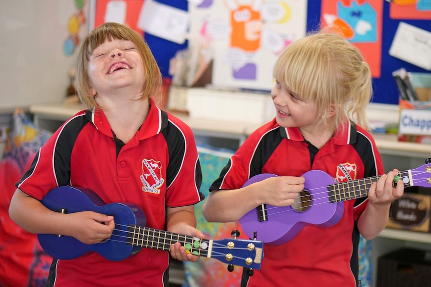Two girls wearing school uniform play ukelele in a classroom, one is giggling.