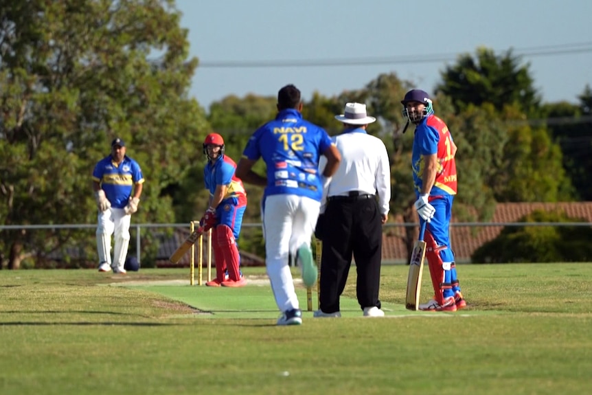 A man runs in to bowl at a cricket game. The batsman, umpire, and other players can be seen too.