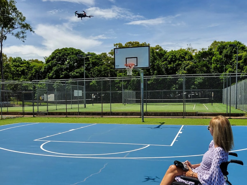A woman in a wheelchair on a blue basketball court operates a drone