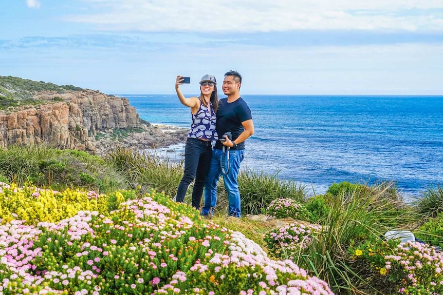 Two people stand on a cliff and one of them holds a phone as they take a selfie with open water behind them.