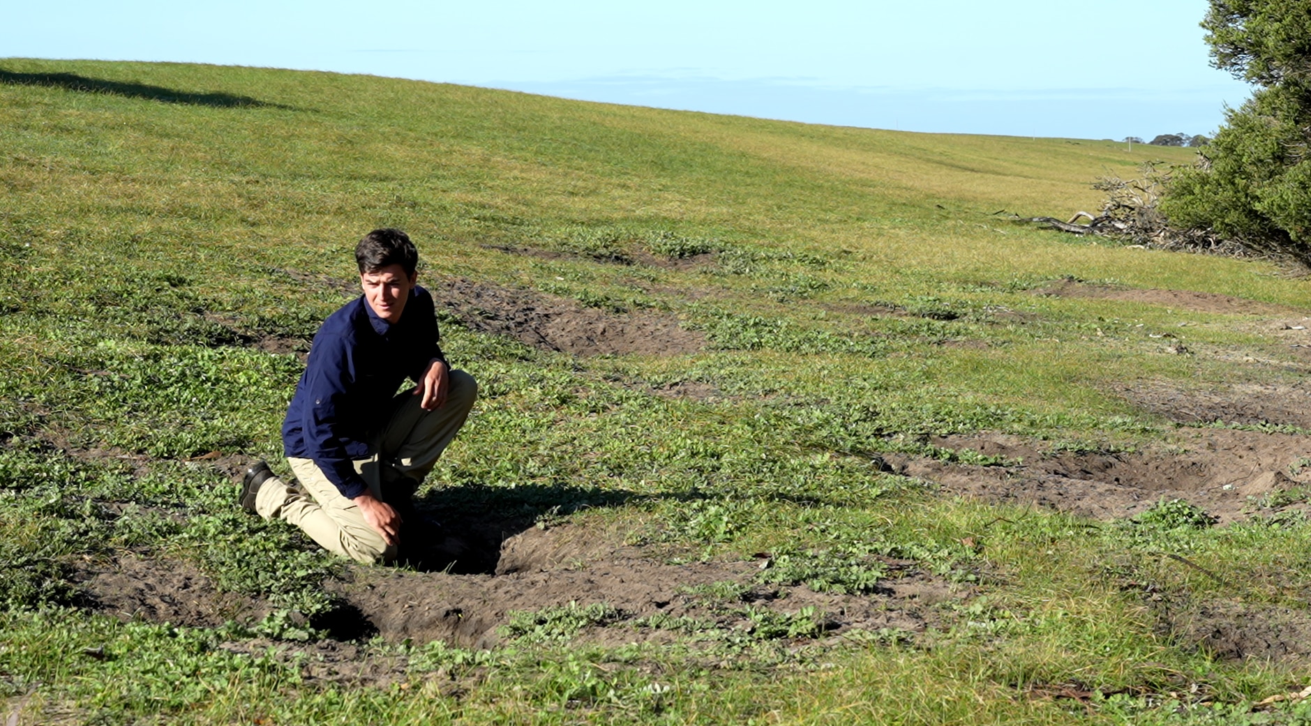 a man in a field kneels next to a hole in the grass.