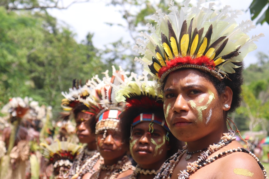 Women wearing traditional clothes including headdresses and beads stand in a line looking past the camera.