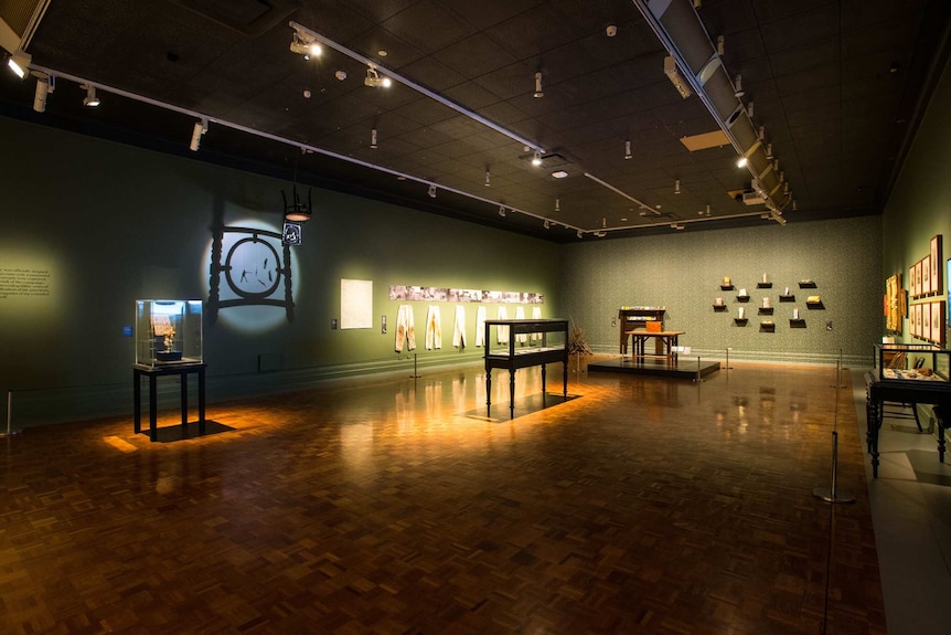 Darkened gallery with parquetry wood floors and eucalyptus-green walls, with several antiques, vitrines and artworks.