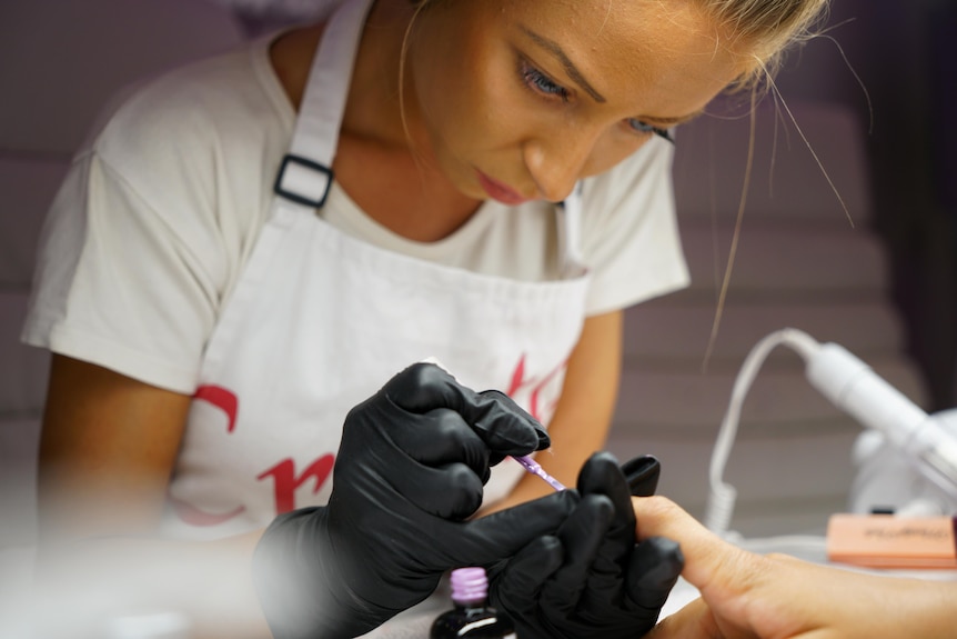 A blonde woman wearing an apron and gloves paints another woman's nails with pink polish