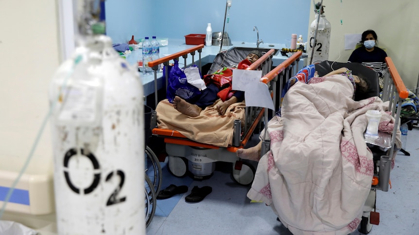 Two patients rest on hospital beds while conected to oxygen tanks, while another person sits behind them