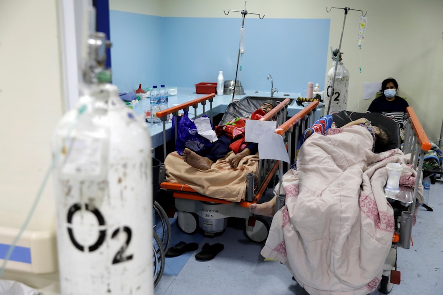 Two patients rest on hospital beds while conected to oxygen tanks, while another person sits behind them