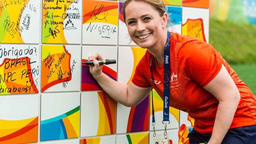 A women signs a wall of signatures with a black marker while turning and smiling at the camera
