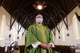 A priest dressed in his green vestments, wearing a mask and standing in an empty church