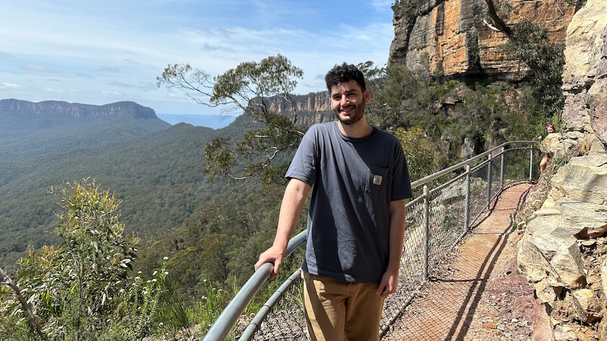 A young man poses for a photo on a cliff face, with mountains behind him.