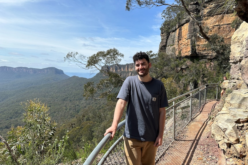 A young man poses for a photo on a cliff face, with mountains behind him.