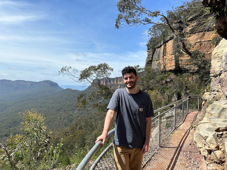 A young man poses for a photo on a cliff face, with mountains behind him.