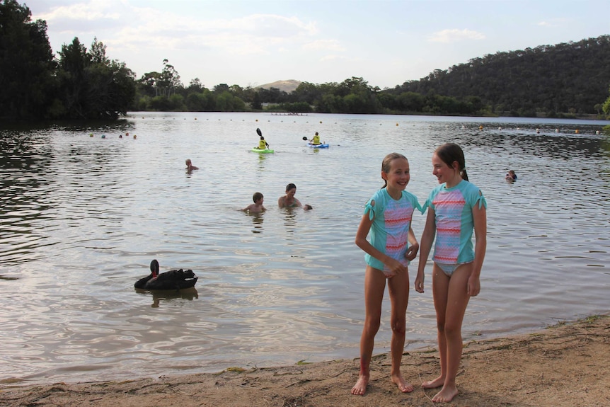 Two girls stand on the shore of Lake Burley Griffin in matching swimmers, there are kayakers and a swan behind them.