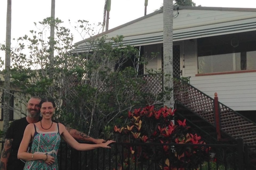 A man stands behind a woman as they rest against a fence with a Queenslander house behind them.