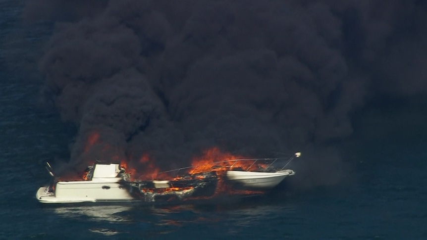 A close-up of a white boat on the water in flames, with black smoke billowing out.