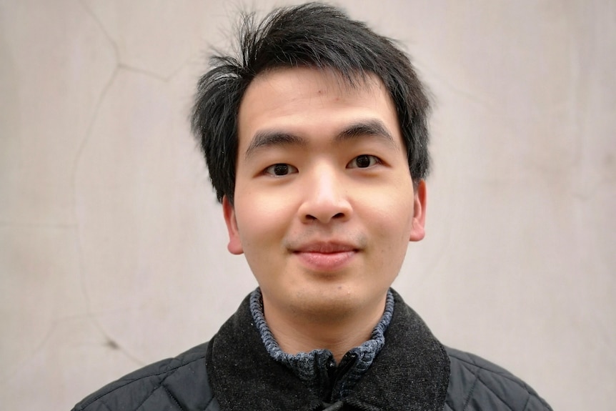 An Asian man with dark hair and black jacket stands against a stone-coloured background