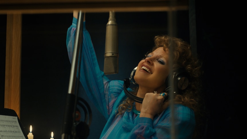 A blonde woman with an 80s perm smiles broadly, headphones held to her ears, as she stands in front of a recording studio mic