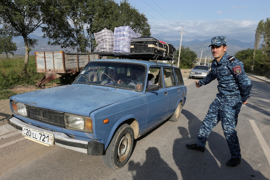 A group of older people drive a battered blue car loaded with luggage past a man in a blue military uniform.