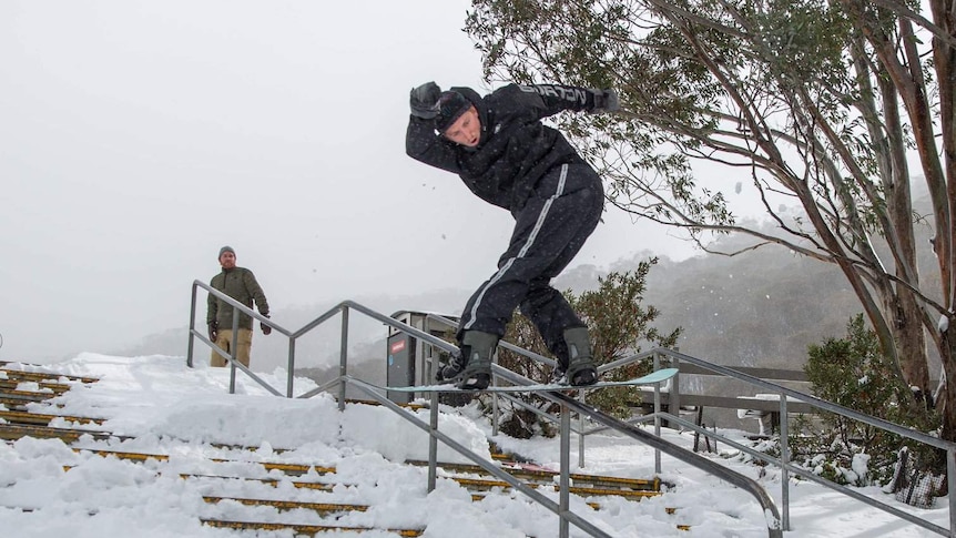 a man on a snowboard skates down a railing on stairs covered with snow with another man watching behind him