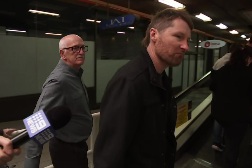 A man with brown hair and a bear speaks to reporters in front of escalators