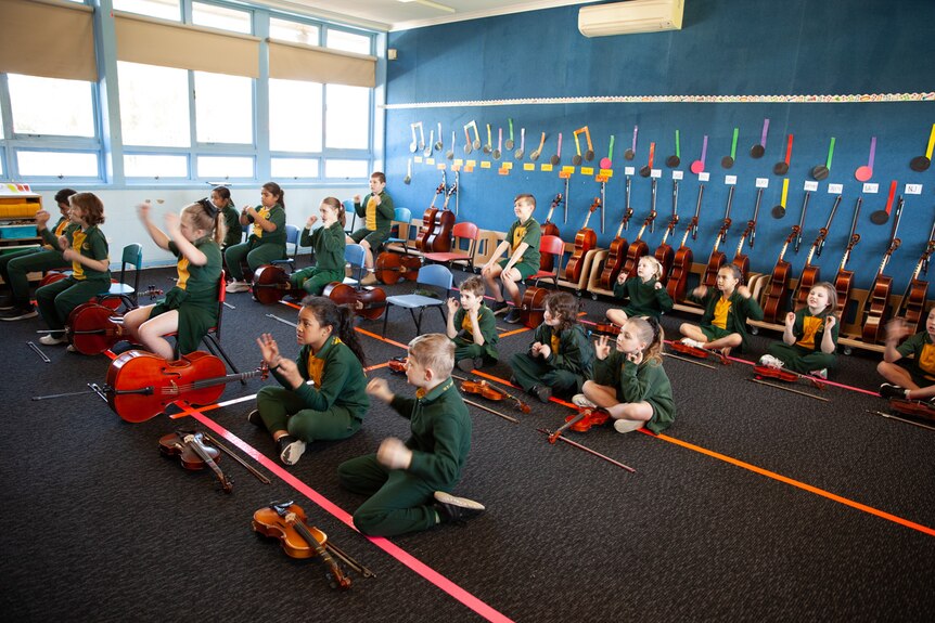 Primary school students in green and yellow uniforms sit cross-legged in a classroom with string instruments in front of them.