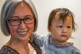 A silver-haired Asian woman with glasses holds an infant in her arms and looks smilingly at the camera.