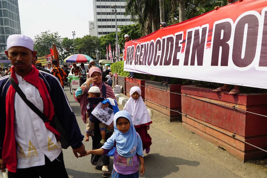 A family is walking down the street and the mother is wiping away a tear. A big protest banner is in the background.