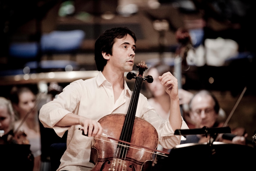 A cellist sits in front of an orchestra. He looks at the conductor. His left hand is raised.