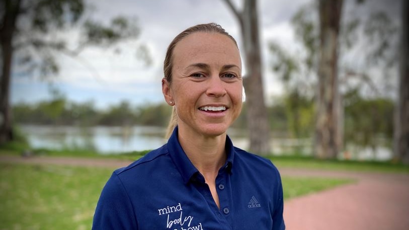 A woman wearing a blue shirt stands in in front of a running track in a park with a river behind her in the background