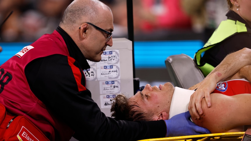 A medic makes contact with an AFL player lying on a motorised stretcher with his neck in a brace after an injury.