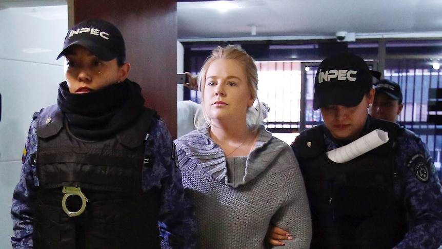 Cassandra Sainsbury wears handcuffs and street clothes and is held by two uniformed guards as she walks through a court building