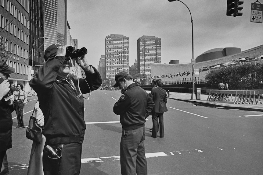 A 1970s black and white photo of an empty city road as people are looking the to the sky with binoculars 