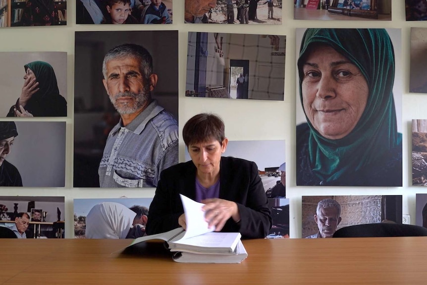 Sahar Francis sits at a desk looking down at a stack of bound papers, with large portraits ofpeople covering the wall behind her