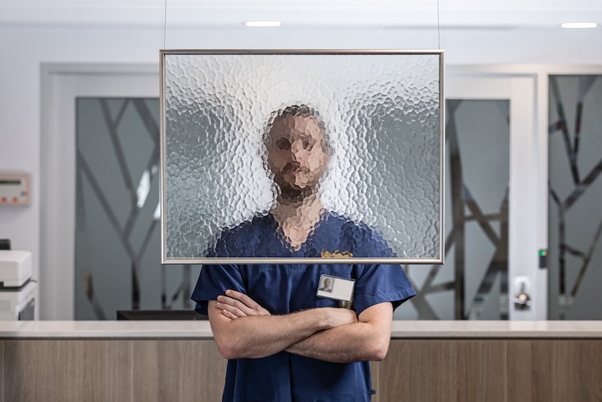 A male nurse in scrubs stands behind a frame of tempered glass which blurs his face.