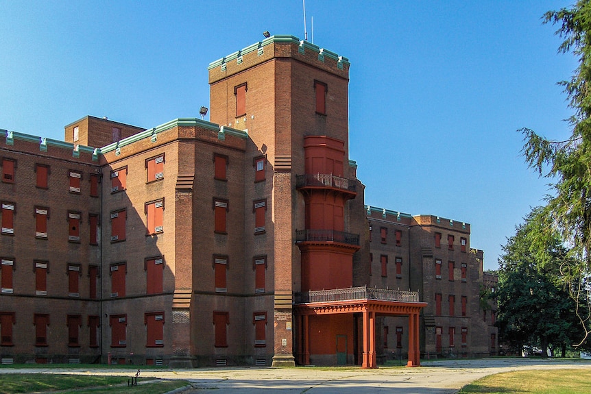 A fortress-like red brick building is seen against a blue sky with a cracked concrete road leading up to it.