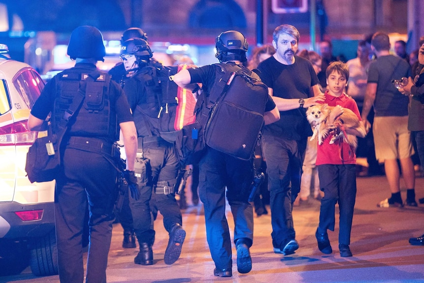 A boy holding a small dog walks, guided by his father, past four heavily clad police.