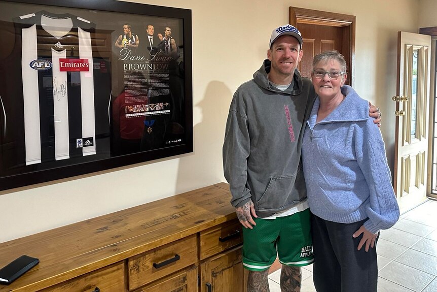 A man in a hoodie and shorts standing with his arm around an older woman standing next to a framed guernsey and plaque
