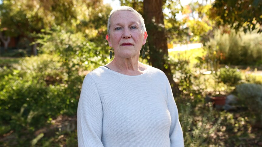 Angela Cooney stands among trees wearing a light coloured jumper and a serious expression