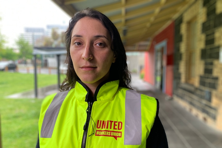 A young woman with long dark hair, wearing a high-vis vest, looks into the camera with a serious expression.