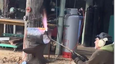 Man kneeling outside on the ground with a welding torch working on a metal helmet for the Ned Kelly movie.