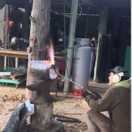 Man kneeling outside on the ground with a welding torch working on a metal helmet for the Ned Kelly movie.
