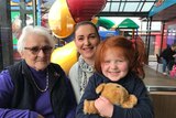 An elderly woman with white hair and glasses is pictured with a young woman and a red-haired girl in a children's playground