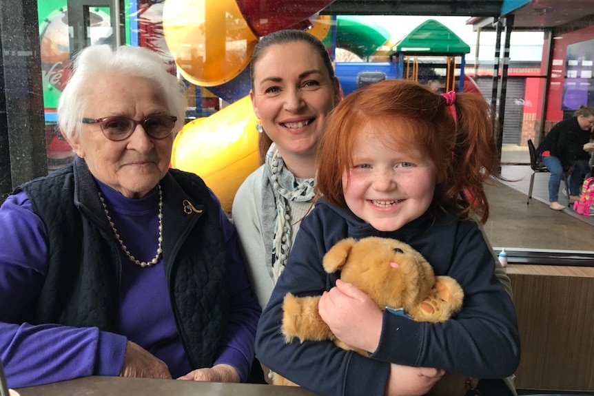An elderly woman with white hair and glasses is pictured with a young woman and a red-haired girl in a children's playground
