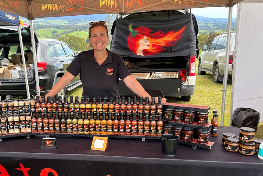 Sue porter wears a black polo with hot chilli mama logo on it, she stands behind her hot sauces with her car in the background