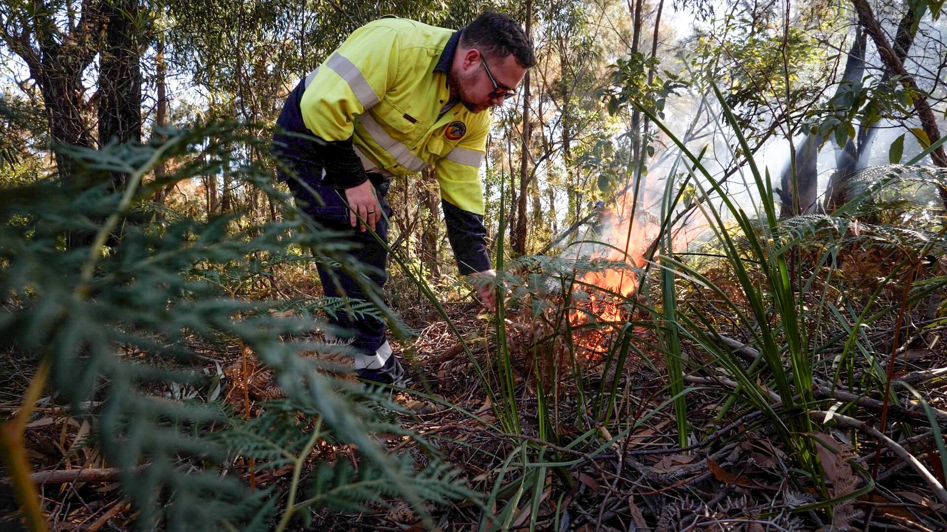 Un hombre se inclina hacia adelante y sostiene helechos ardiendo en la base de un trozo de hierba seca.