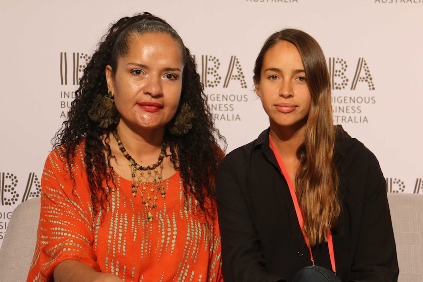 Two Indigenous women sitting on a couch in front of a background poster saying Indigenous Business Australia