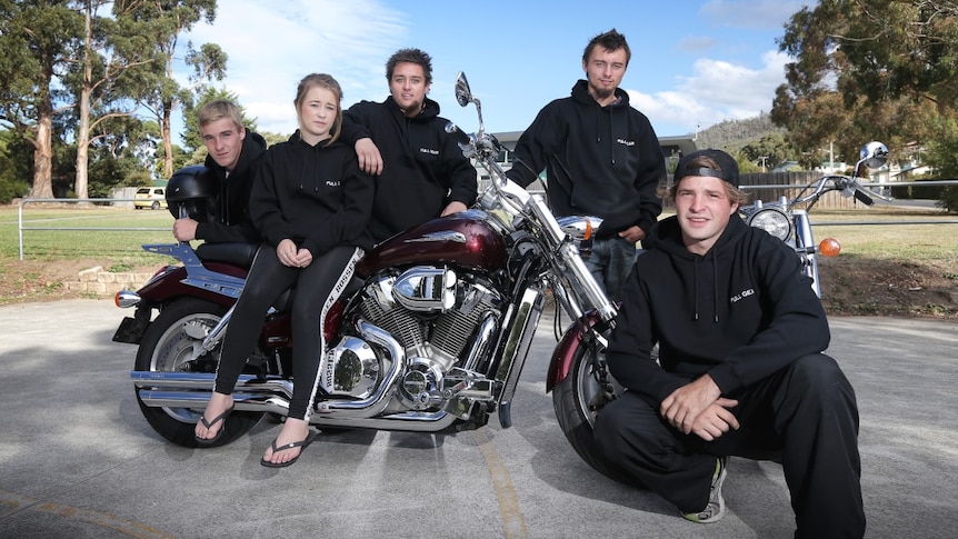 A group of young people dressed in black posing for a photo around a big black motorbike.