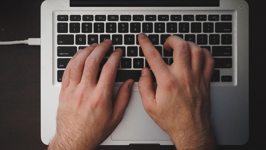 Close-up shot of two hands with dark-ish hair on them on the keyboard of a silver laptop.