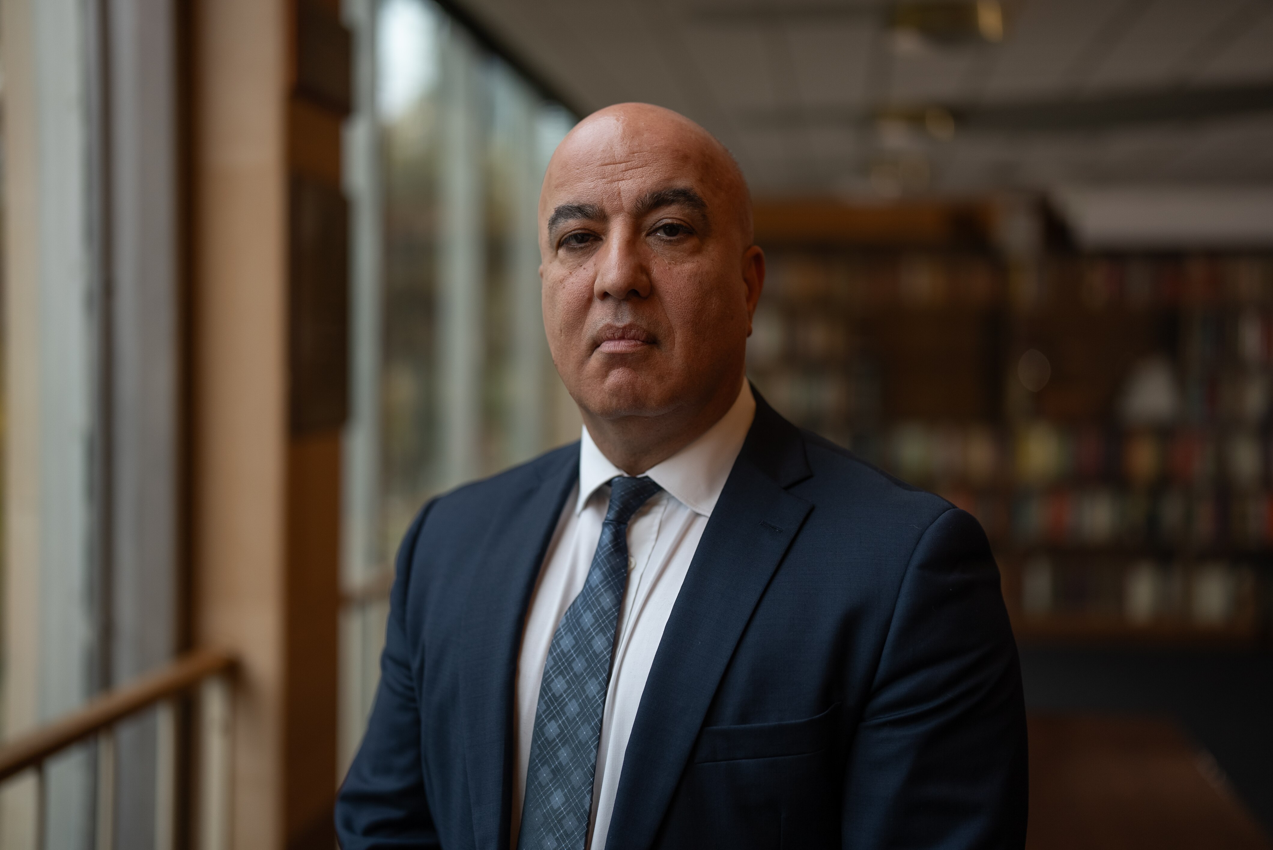 A man in a suit, standing in a room with windows and a large bookcase looks at the camera with a serious expression.